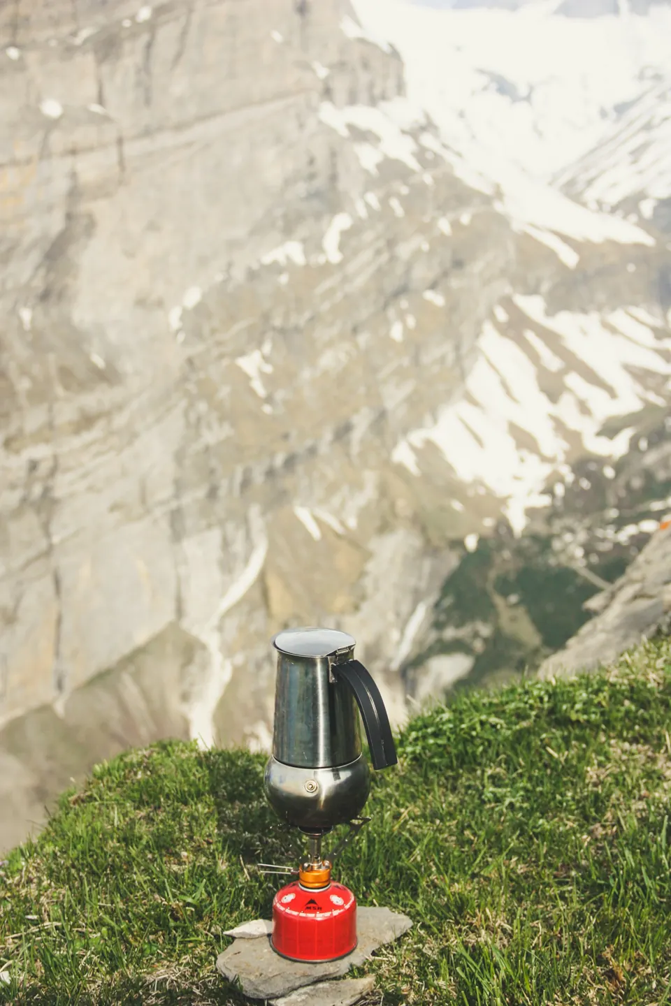 A metal kettle on a small canister stove outdoors below a mountain cliff.